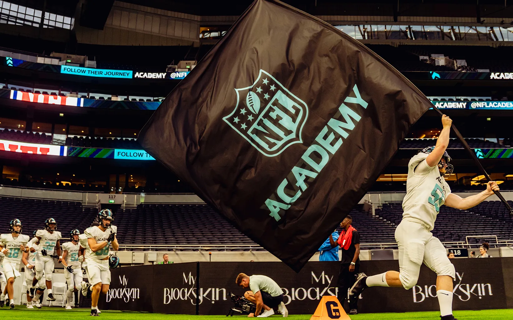 Ein Spieler der NFL Academy läuft mit einer großen schwarzen Flagge mit dem Logo der „NFL Academy“ über das Spielfeld, während sein Team im Hintergrund das Stadion betritt.